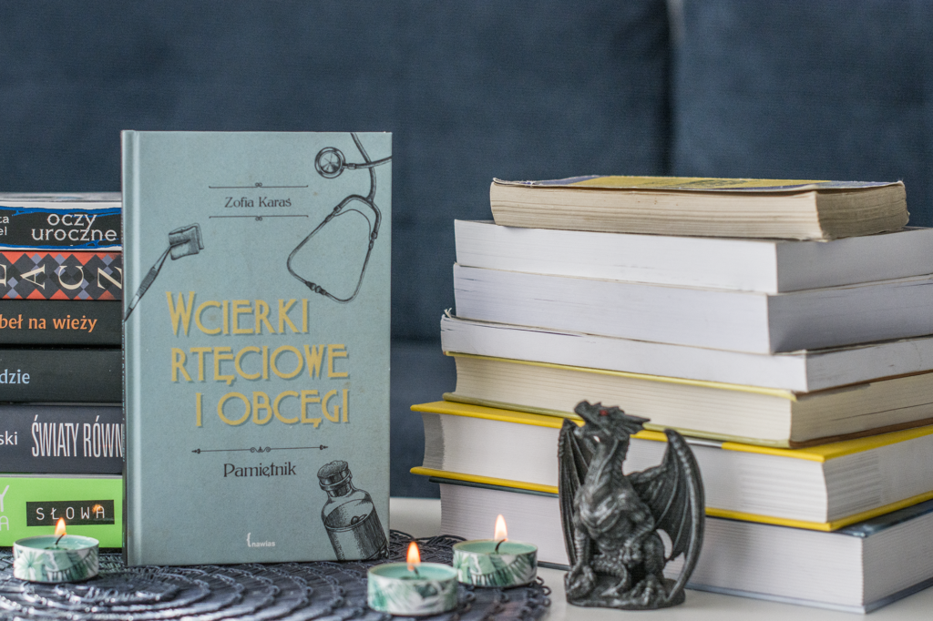 Wcierki rtęciowe i obcęgi Zofia Karaś - książka stoi na placie na podkładce, przed nią świeczki, nieco po prawej figurka smoka, otaczają ją inne książki, w tle niebieska kanapa  - ilustracja do recenzji pamiętnika polskiej lekarki