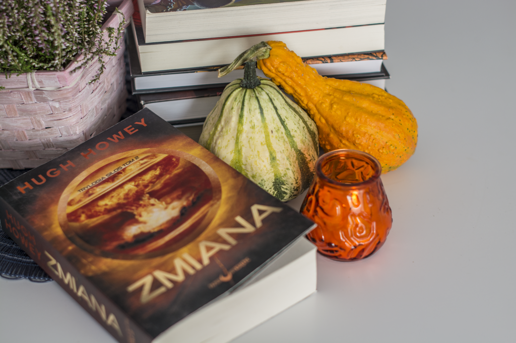 Zmiana Hugh Howey - książka lezy na blacie w otoczeniu ozdobnych dynii i świeczki. W tle wrzos w koszyku i książki - ilustracja recenzji powieści post-apokaliptycznej