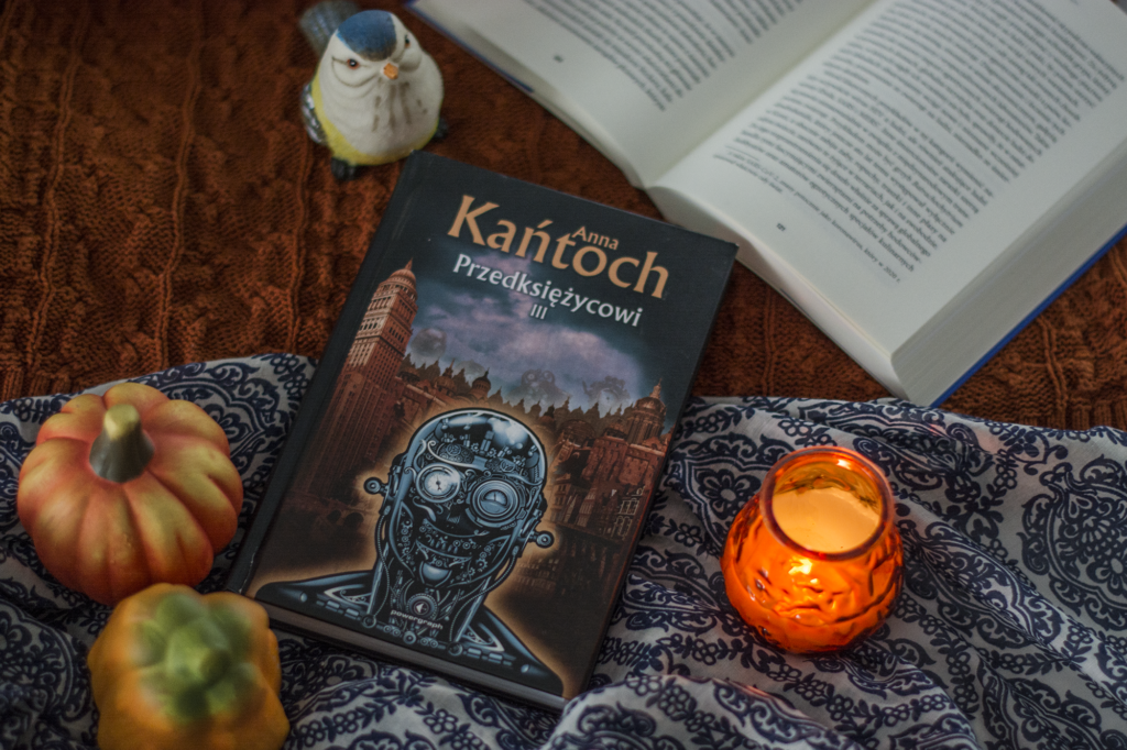 Przedksiężycowi. Tom III Anna Kańtoch - książka leży na tkaninach, w tle otwarta inna książka oraz ozdoby: dynia, ceramiczny ptaszek i świeczka - recenzja polskiej ksiązki steampunkowej