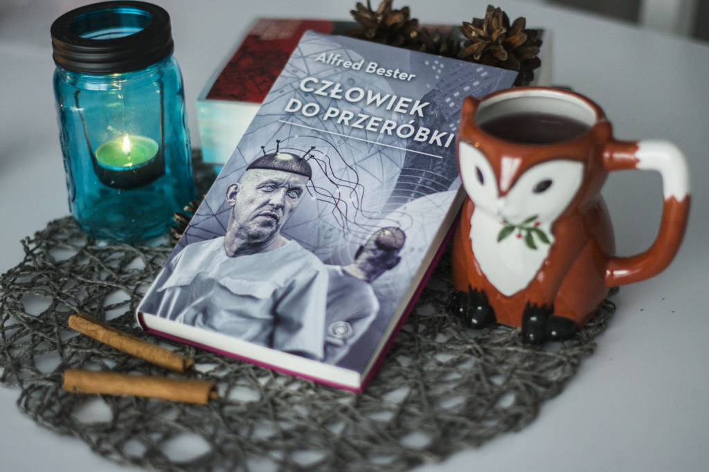Człowiek do przeróbki Alfred Bester - książka leży na blacie, podpierając się o inną książkę. Ozdoby wokół: kubek-zwierzątko, lampion, szyszki, cynamon - ilustracja do recenzji powieści cyberpunkowej