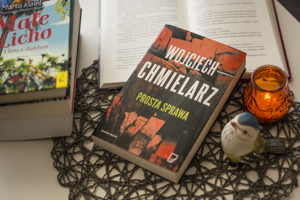 Prosta sprawa Wojciech Chmielarz - książka leży na blaccie w otoczeniu książek, świeczki oraz figurki ptaszka - ilustracja do recenzji polskiej zekranicowanej powieści sensacyjnej
