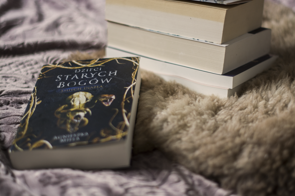Śmiech diabła Agnieszki Mieli - książka leży na kocu, obok inne książki - ilustracja do recenzji powieści high fantasy
