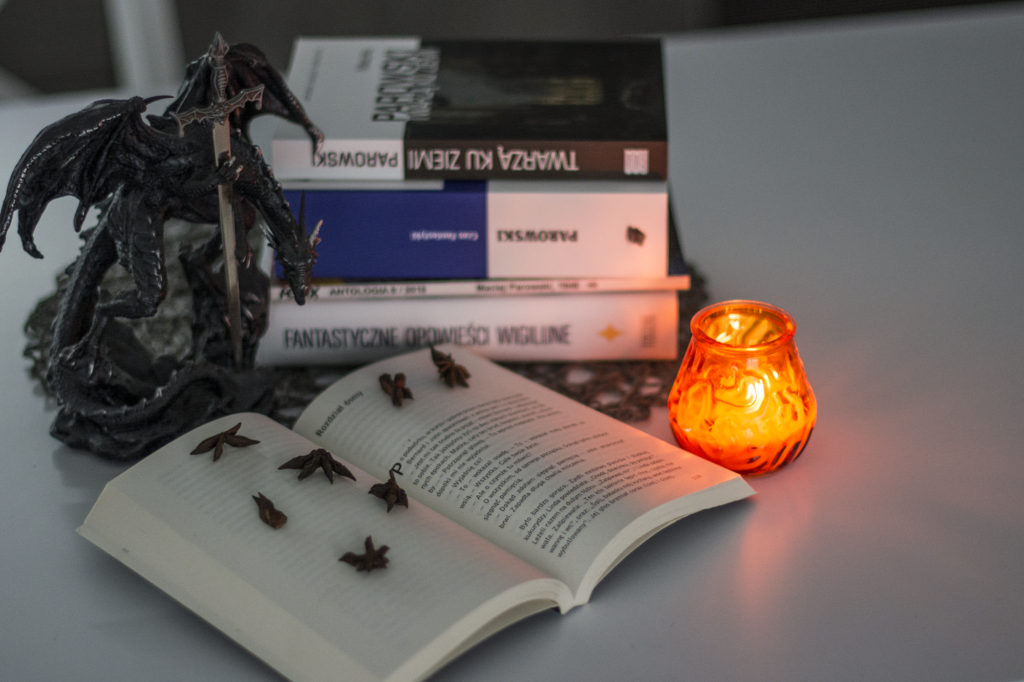 Nowy wspaniały świat Adolus Huxley  - książka leży otwarta na blacie w otoczeniu ozdób: figurki smoka, goździków, świeczki, innych książek - ilustracje do recenzji dystopii, fantastyki socjologicznej