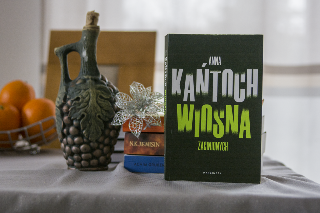 Wiosna zaginionych Anna Kańtoch - książka stoi na stole, w tle inne książki, gliniana butelka, pomarańcze - ilustracja do recenzji polskiego kryminału