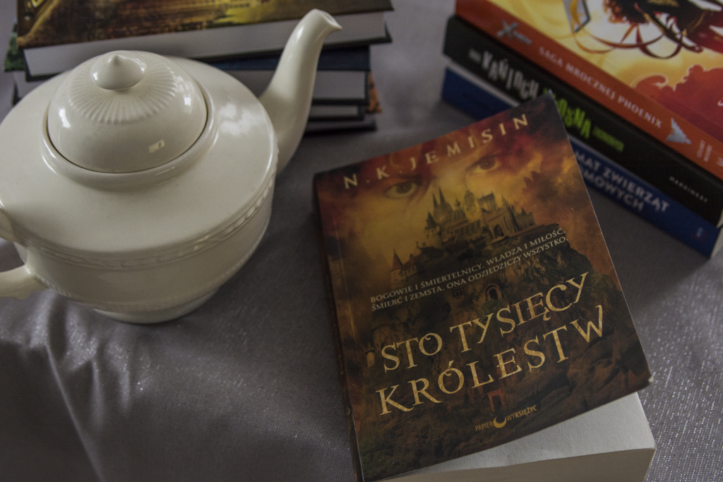 Sto tysięcy królestw Jemisin - książka lezy na blacie w otoczeniu książek i porcelanowego czajnika - ilustracja do recenzji